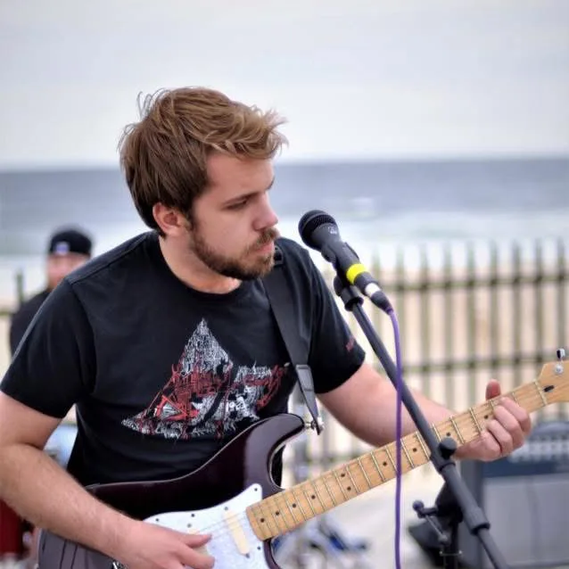 Ryan playing a red Stratocaster into a microphone on a boardwalk by the sea.